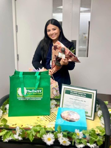 Ashley Espinoza in nursing scrubs holding flowers behind a award plaque and trophy